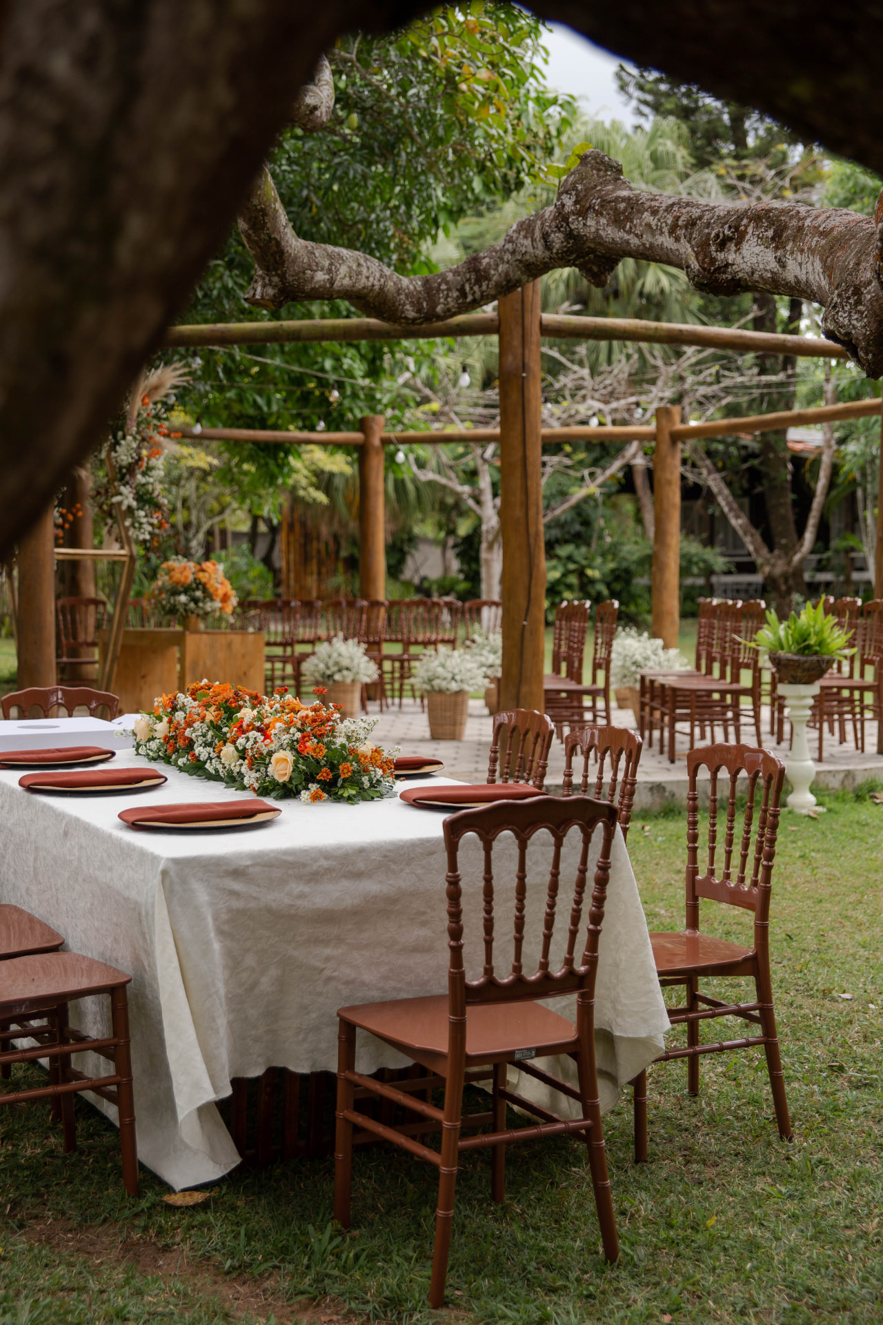 Mesa dos noivos posta no gramado com arranjo floral e cadeiras de madeira